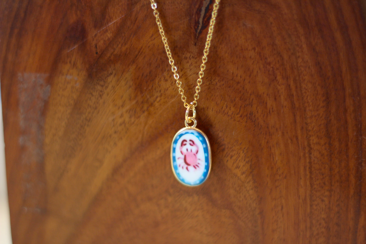 A gold chain necklace with an oval pendant featuring a colorful illustration of a crab on a blue and white background, displayed on a wooden surface.