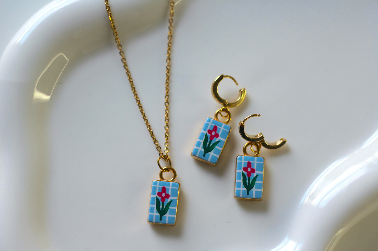 A gold jewelry set featuring a necklace and matching hoop earrings with rectangular charms decorated with a blue grid pattern, pink flowers, and green leaves, displayed on a white ceramic dish.