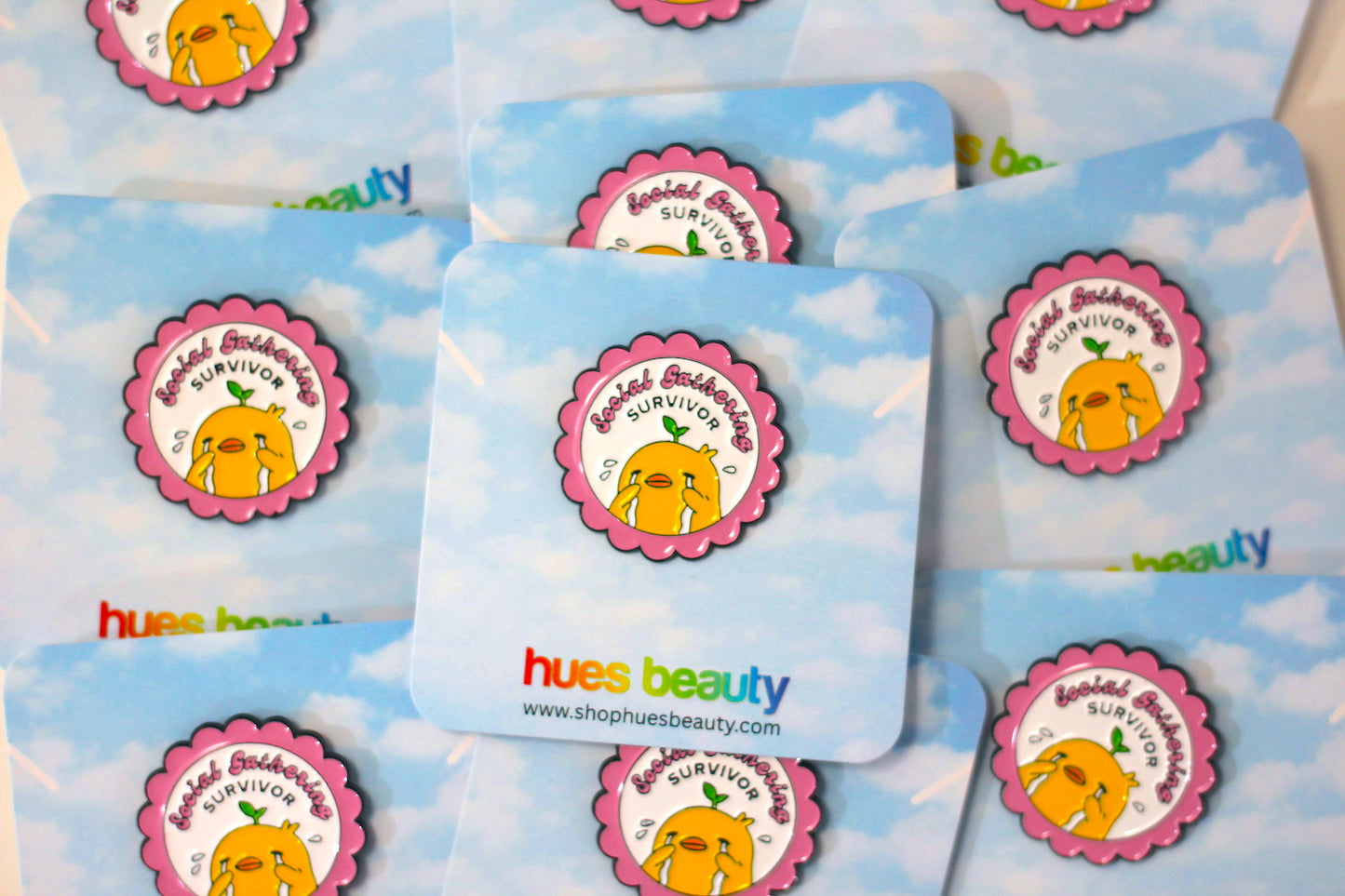 A close-up of multiple round enamel pins arranged in a fan-like layout. Each sticker has a light blue sky background with white clouds and a circular character sticker in the center featuring a yellow duck inside a pink flower frame and the words "social gathering survivor".