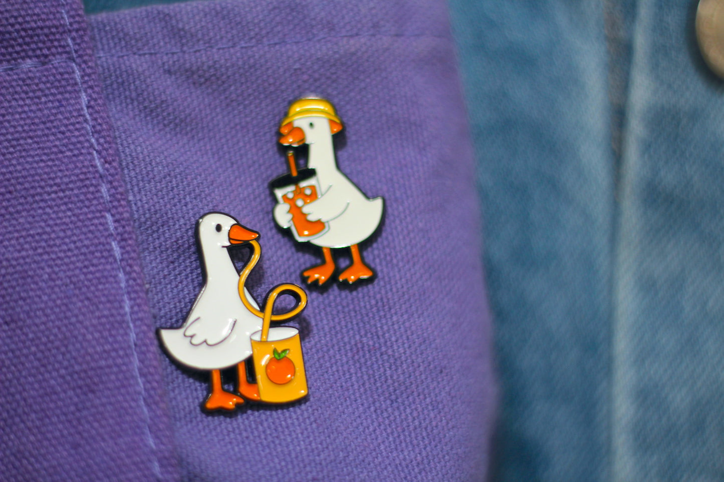 Two small duck enamel pins displayed on a purple fabric pocket next to a blue denim jacket.