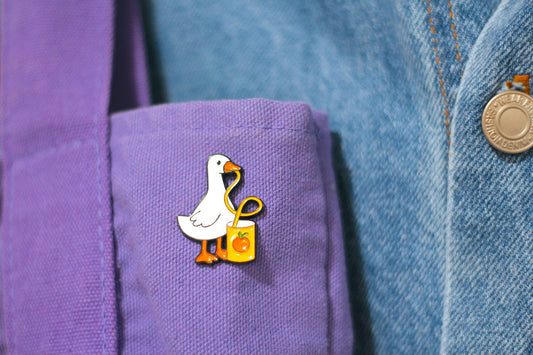A close-up of a white goose enamel pin with orange feet and a yellow bag, pinned to a purple shirt cuff next to a blue denim jacket.