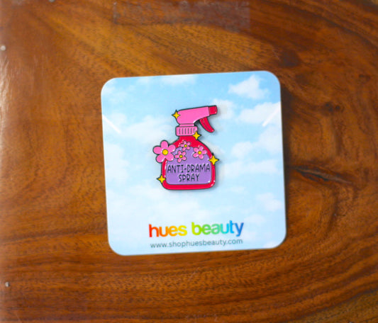 A small enamel pin shaped like a pink spray bottle with flowers and the words 'ANTI-DRAMA SPRAY' on a blue sky printed backing card, placed on a wooden surface. The card shows the brand name 'hues beauty'.