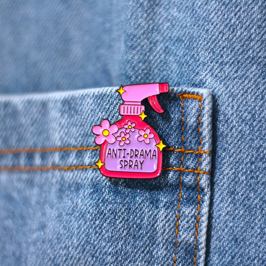 A pink enamel pin shaped like a spray bottle with flowers and the text 'ANTI-DRAMA SPRAY' pinned onto blue denim fabric.