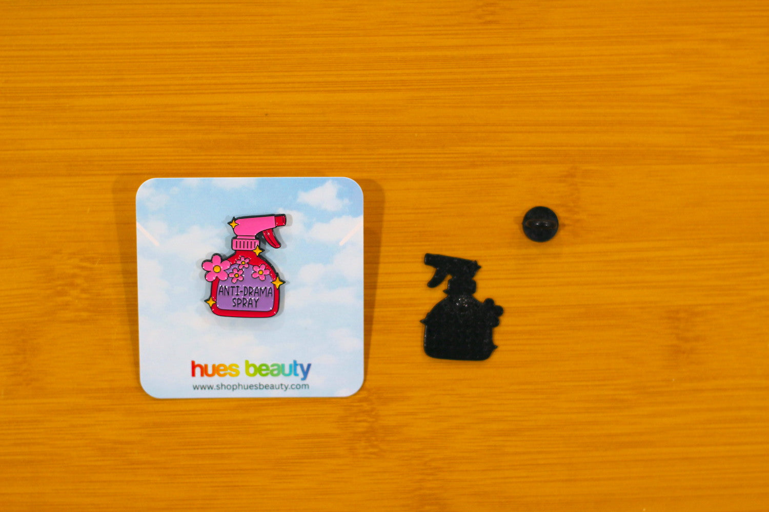A pink and purple enamel pin shaped like a spray bottle with the words 'ANTI-DAMAGE PROPHYLAXIS' is displayed on its blue backing card next to the black metal pin and a small black button, all on a wooden surface. The brand 'hues beauty' is visible on the card.