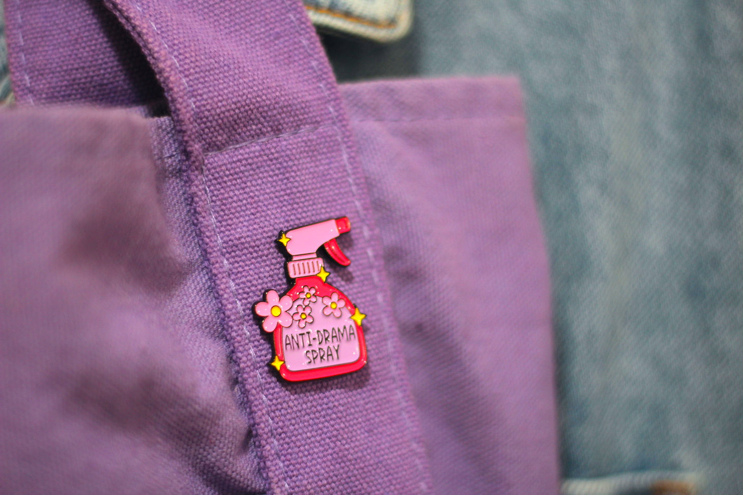 A close-up of a purple canvas tote bag featuring a pink enamel pin shaped like a spray bottle labeled 'ANTI-DRAGON SPRAY' with yellow flowers and a red trigger sprayer, set against a blurred blue denim background.
