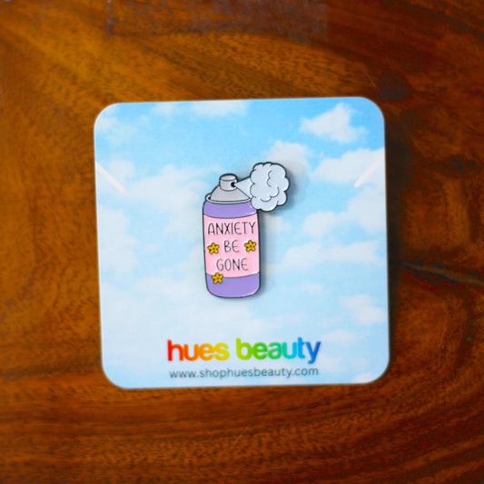 An enamel pin shaped like a lavender spray can with the words 'ANXIETY BE GONE' and small yellow flowers, mounted on a square blue backing card that reads 'hues beauty' and is photographed on a wooden surface.
