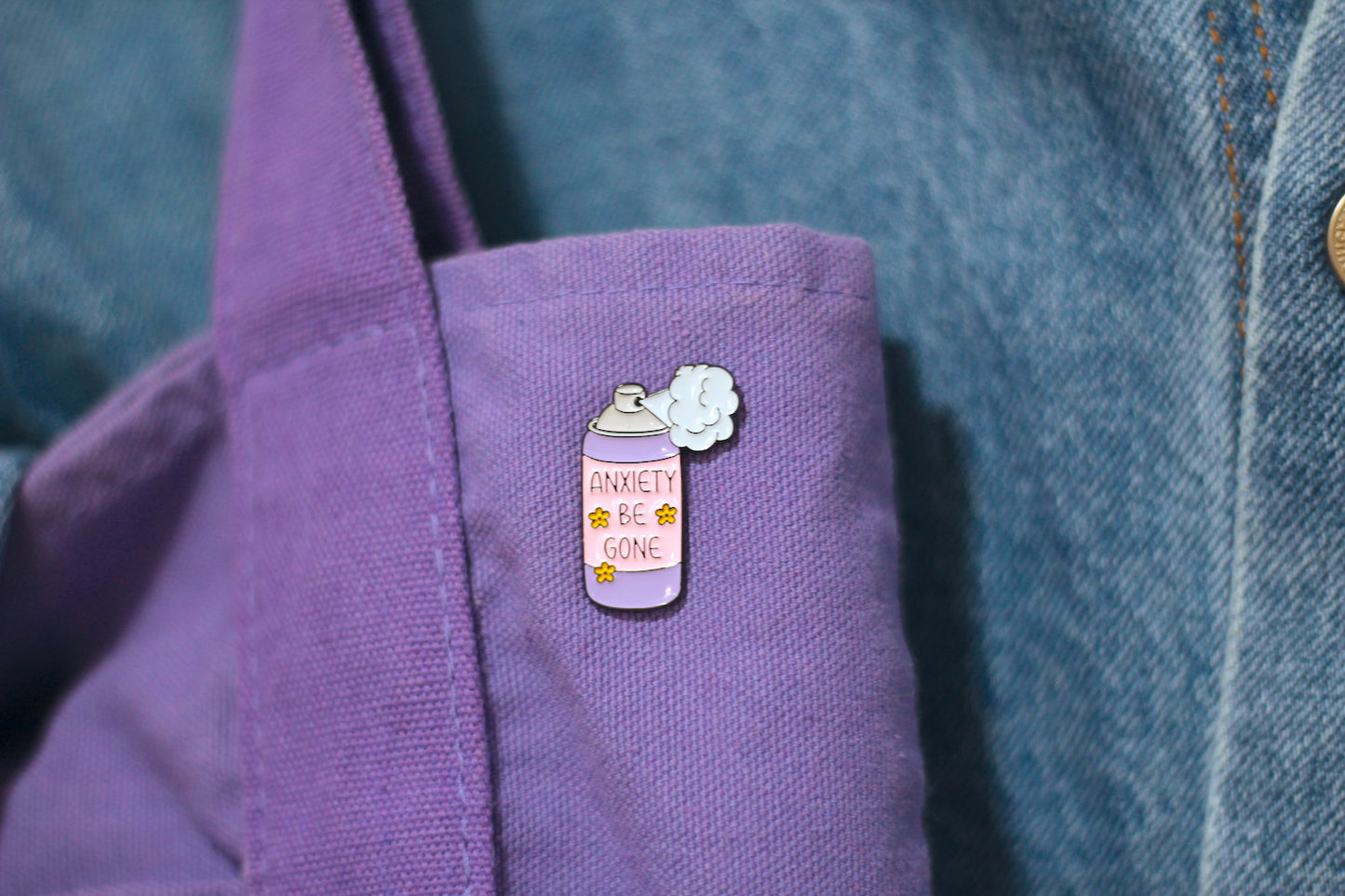 A close-up of a lavender enamel pin shaped like a spray can with the text 'ANXIETY BE GONE' and a small white flower, pinned onto a purple fabric surface next to a blue denim jacket.