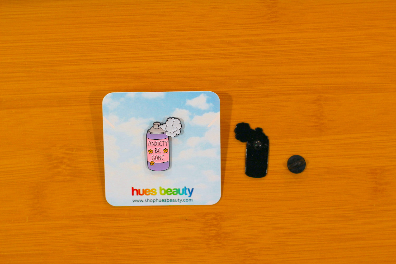 An enamel pin shaped like a coffee cup with the words 'anxiety be gone' on a light blue cloud-patterned backing card, displayed on a wooden surface next to its black backing and a small round pin.
