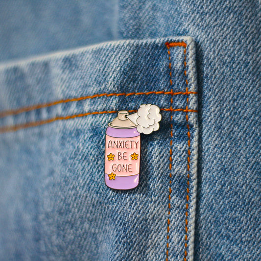 A close-up of a lavender spray-can enamel pin with the words 'ANXIETY BE GONE' and small yellow stars, pinned to a blue denim pocket.