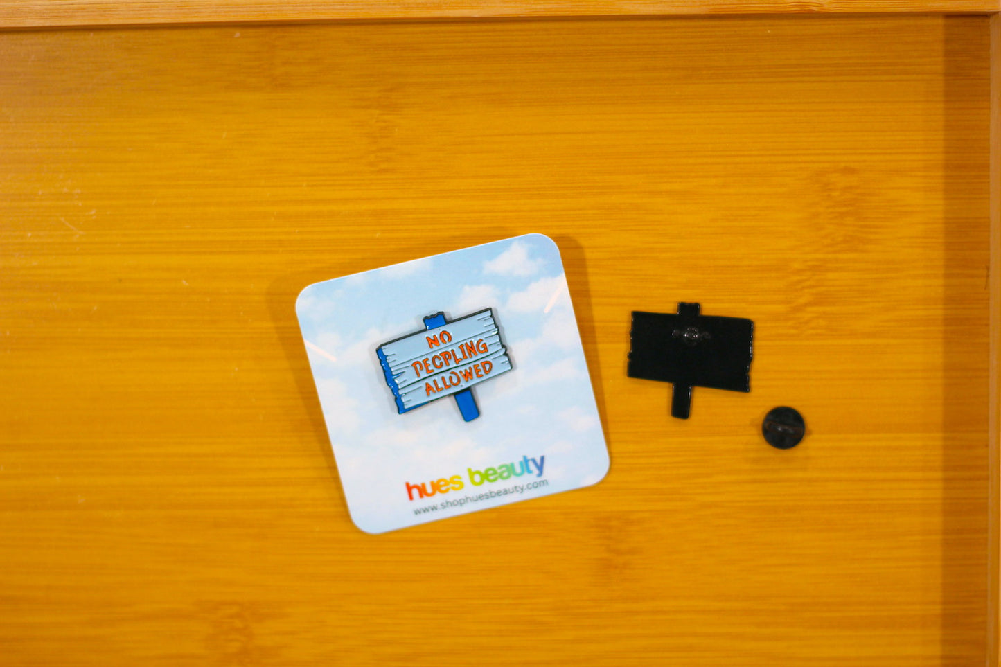 A square white card with rounded corners featuring a blue sky and clouds background, a sign that reads 'NO PEOPLES ALLOWED' in red, and the multicolor 'hues beauty' logo at the bottom, shown on a wooden surface next to a small black accessory.