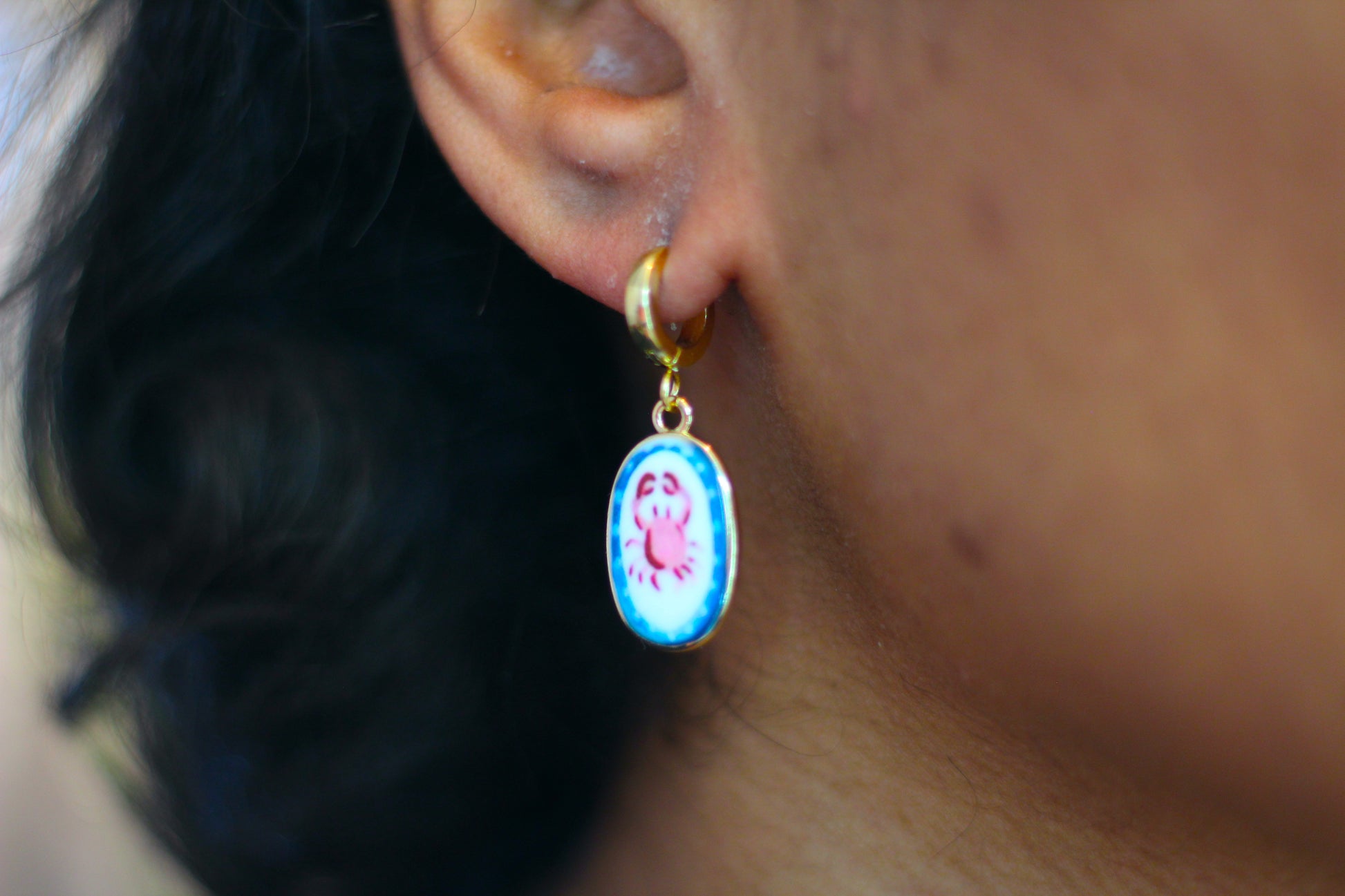 A close-up of a person's ear wearing a gold hoop earring with an oval dangling charm featuring a crab design on a light blue background.