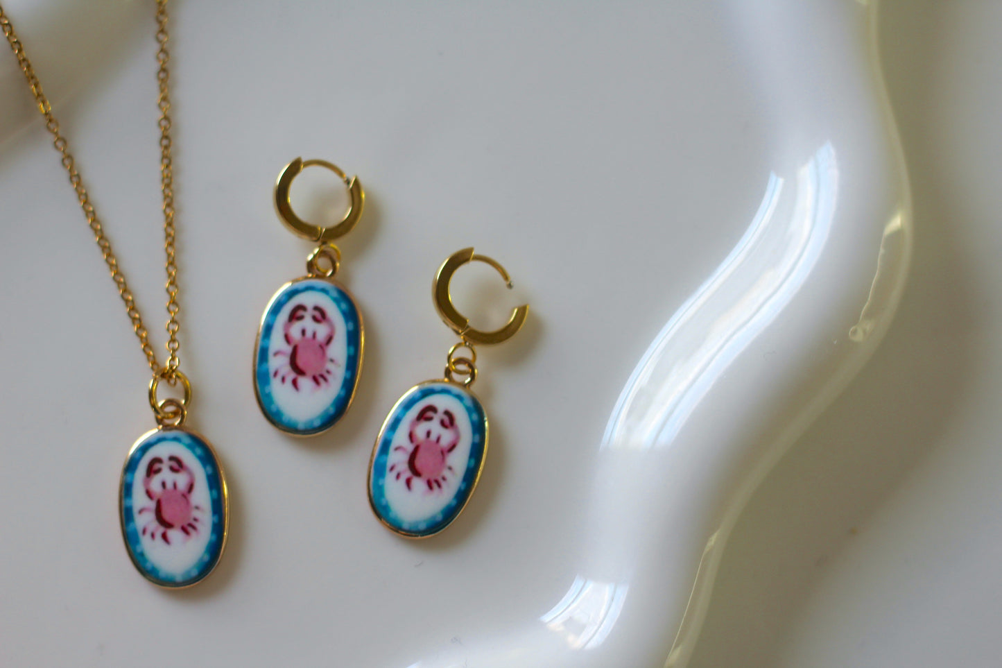 Gold jewelry set featuring a necklace and two matching drop earrings with oval enamel pincushion-style crab charms, displayed on a white ceramic dish.