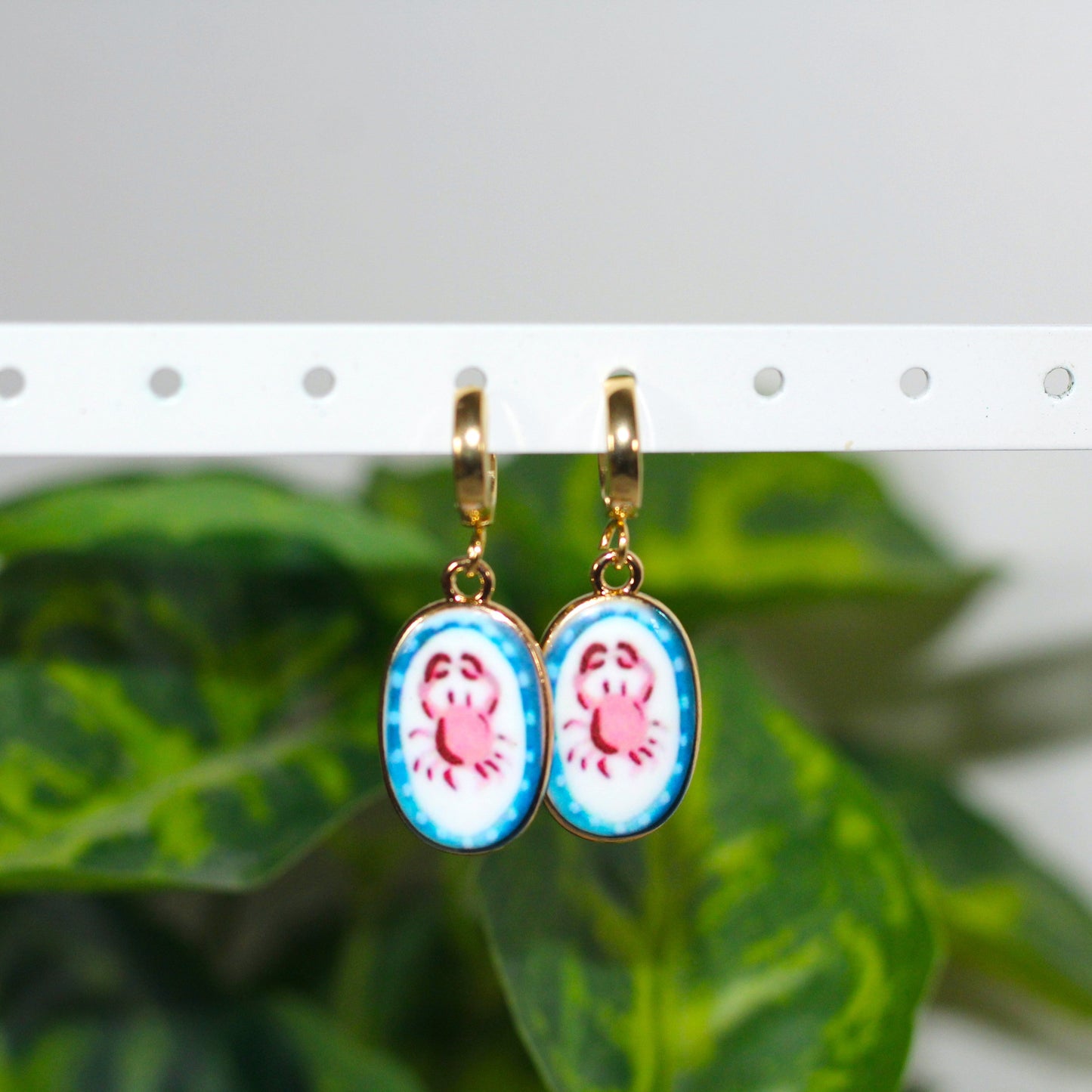 A pair of oval drop earrings with gold hooks and blue bezel settings, featuring a crab design on a white background with light blue accents, hanging in front of a blurred green plant.