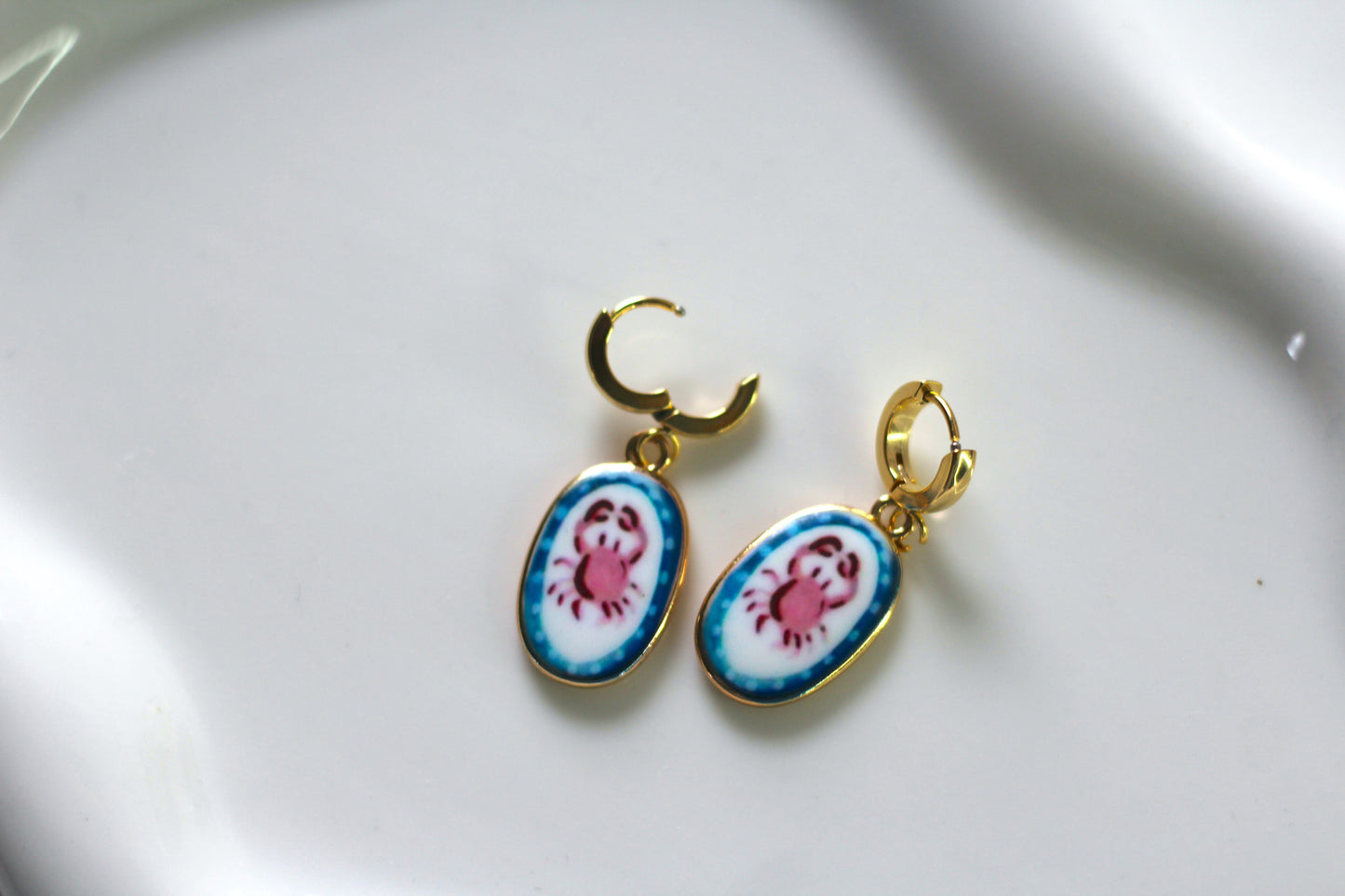 A pair of gold hoop earrings with oval enamel charms featuring a pink crab design on a white and blue background, displayed on a white ceramic surface.