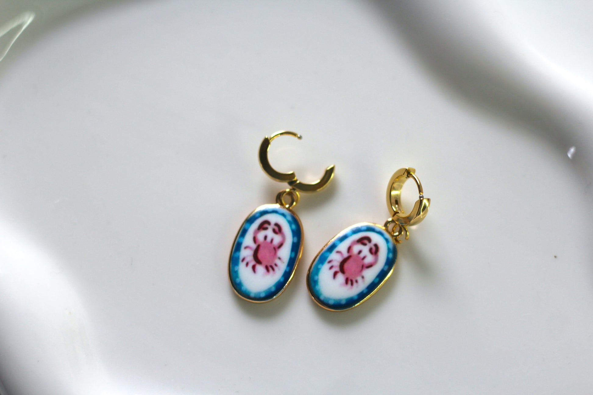 A pair of gold hoop earrings with oval enamel charms featuring a pink crab design on a white and blue background, displayed on a white ceramic surface.
