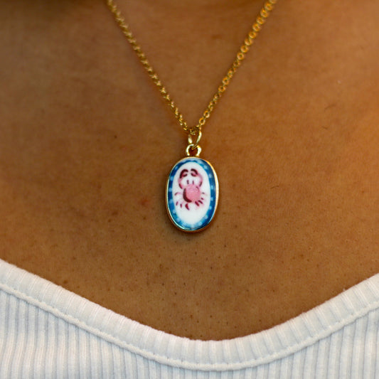 A gold chain necklace with an oval pendant featuring a crab drawing on a white and blue background, worn on a person's neck over a white ribbed top.