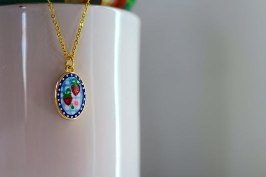 A gold chain necklace with an oval pendant featuring a colorful illustration of strawberries on a blue and white background, hanging against a white cylindrical surface.