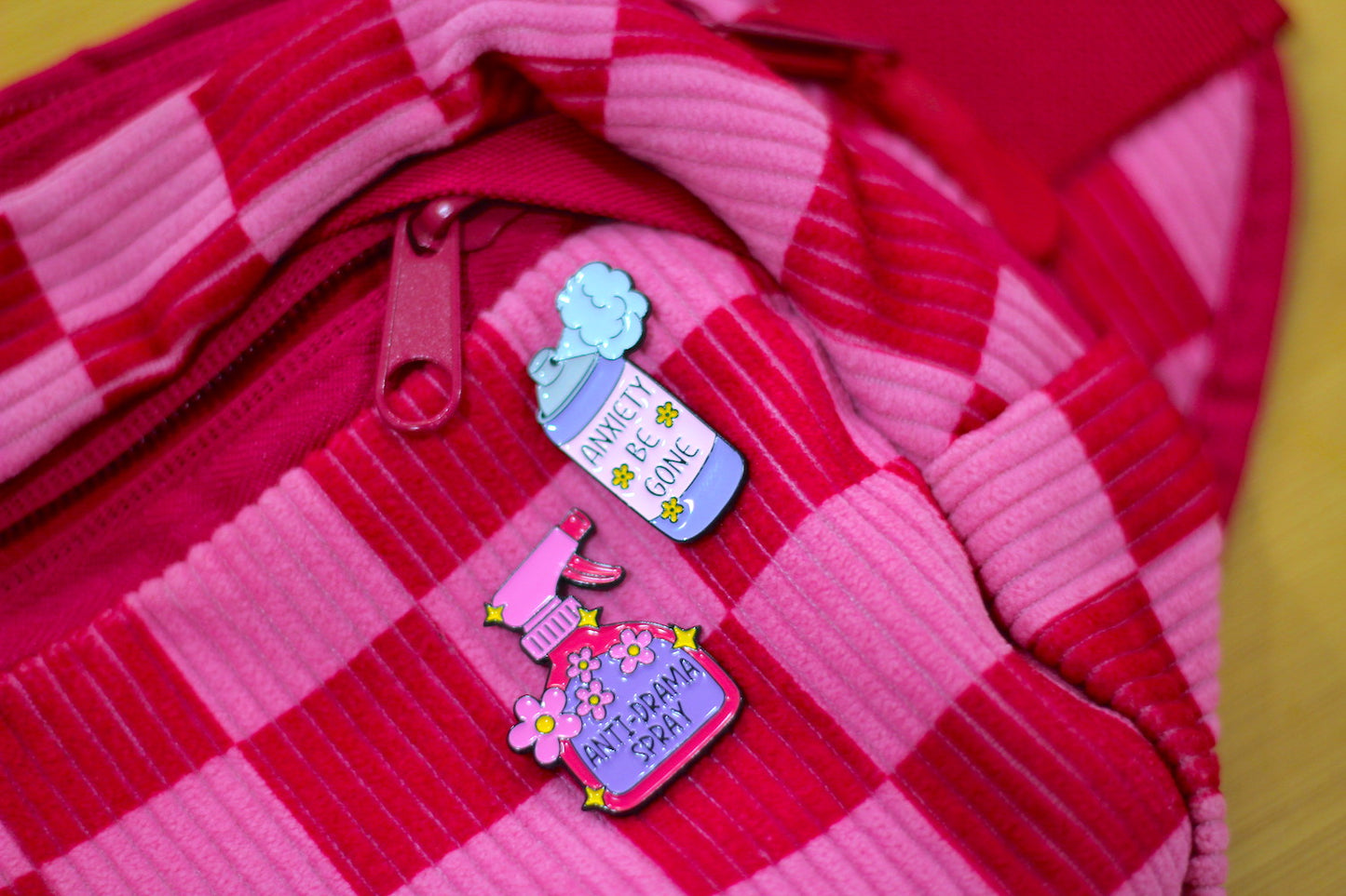 A close-up of a pink and red checkered zippered pouch or bag, featuring two small enamel pins attached near the zipper. The pins depict a blue bottle labeled 'WATERING CANVAS' and a pink spray bottle labeled 'MY SPRAY SCENE' with flowers.