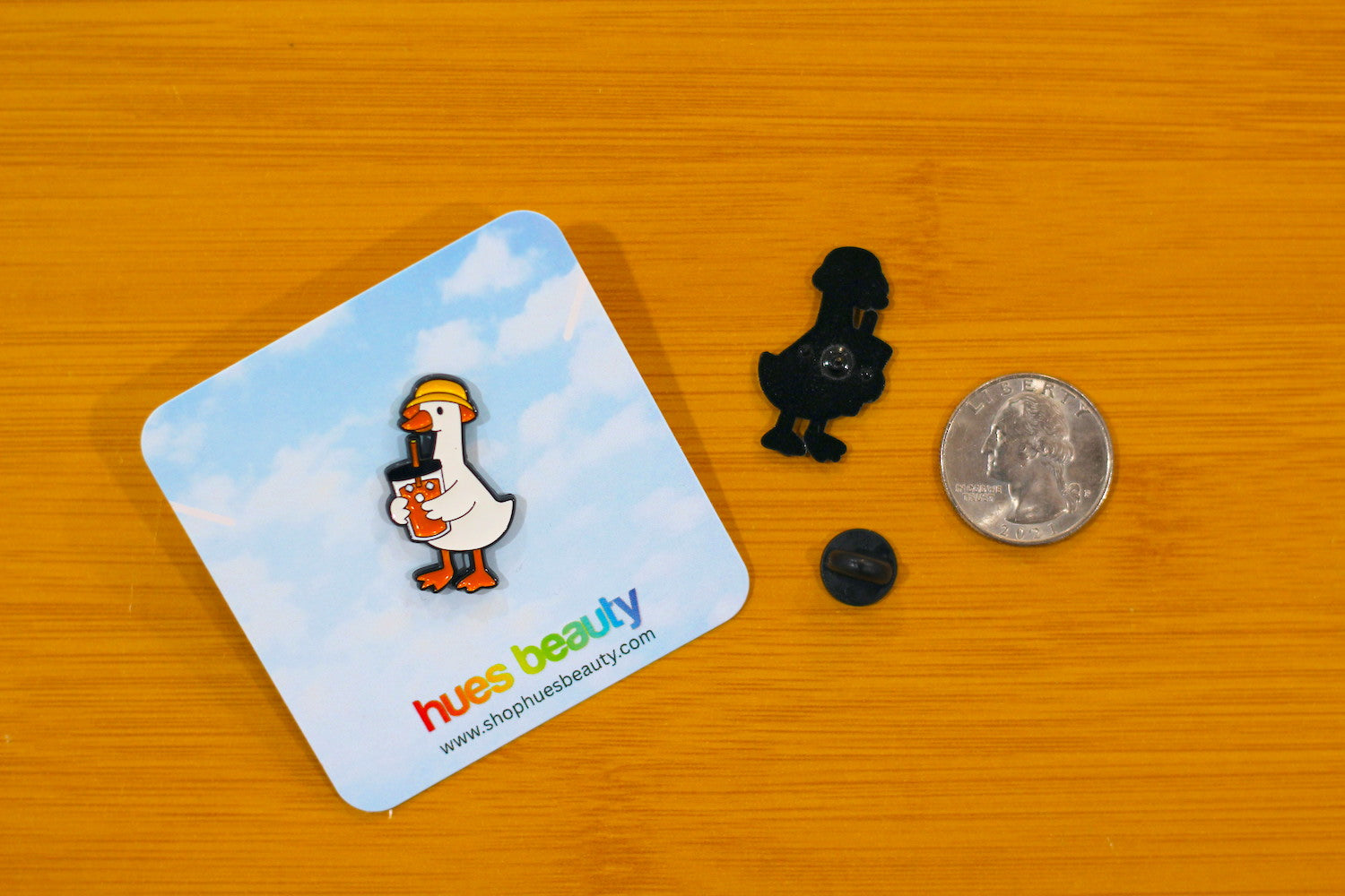 A small enamel pin shaped like a duckling is displayed on a wooden surface next to its branded backing card, a black dome clasp, and a coin for scale. The backing card features a cartoon duck holding a megaphone against a blue sky background, with the brand name 'hues beauty' printed at the bottom.