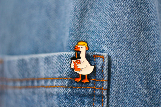 A small enamel pin shaped like a white duck wearing a yellow hard hat and holding an orange drink, attached to a blue denim pocket.