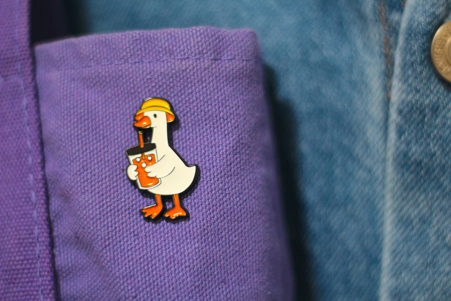 A small enamel pin shaped like a white goose wearing a yellow hard hat and holding an orange drink, attached to a purple fabric cuff next to a blue denim jacket.