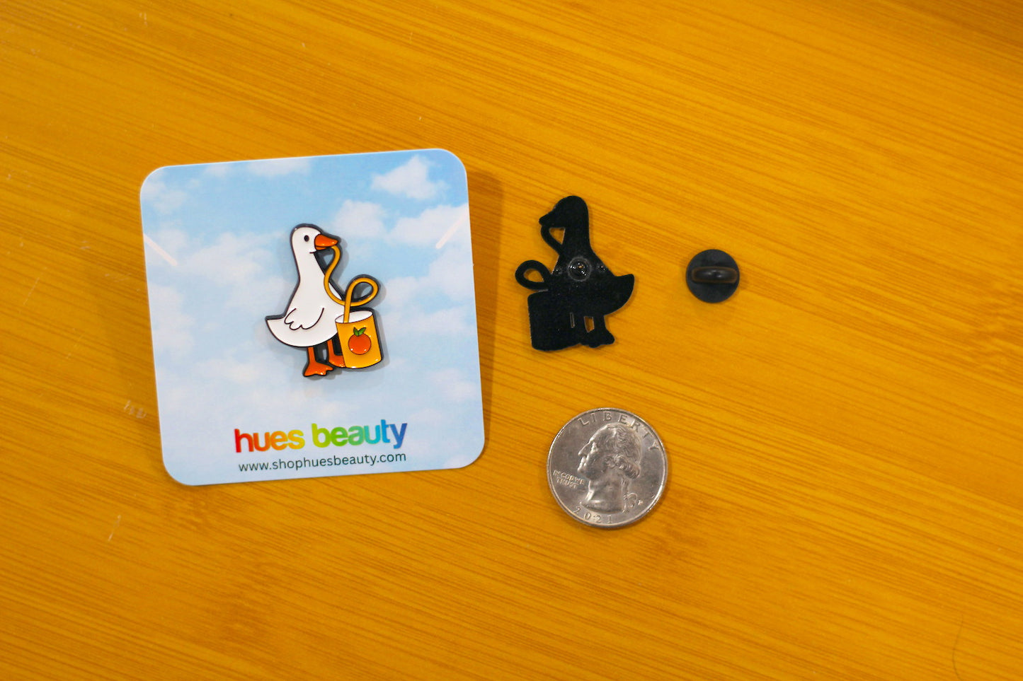 An enamel pin of a white duck with an orange beak and a yellow-orange bag, displayed on its blue backing card next to a black clip and a US quarter for scale, on a wooden surface.