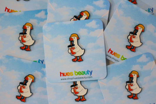 Several square cards with a blue sky and white cloud print, each featuring a cartoon duck wearing a yellow hat and holding a black camera. The center card shows the text 'hues beauty' and a website URL.