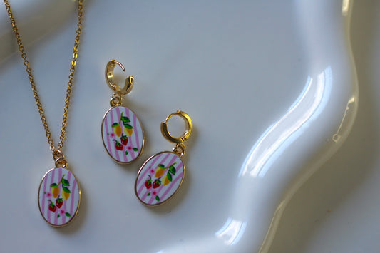 Gold-tone jewelry set featuring a necklace and matching drop earrings with oval charms decorated with pink stripes and a colorful floral fruit design, displayed on a white ceramic dish.