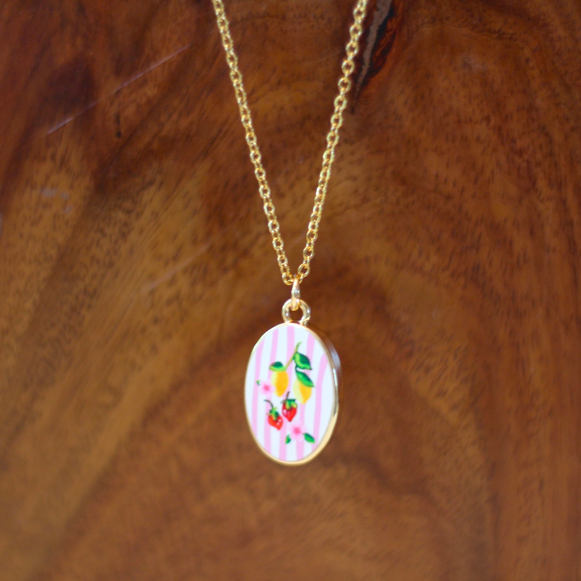 A gold chain necklace with an oval pendant featuring a colorful floral pattern on a light pink background, displayed against a dark wood grain background.