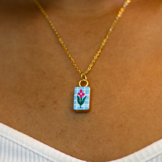 A close-up of a person wearing a delicate gold chain necklace with a small rectangular pendant featuring a colorful floral design on a light blue background.