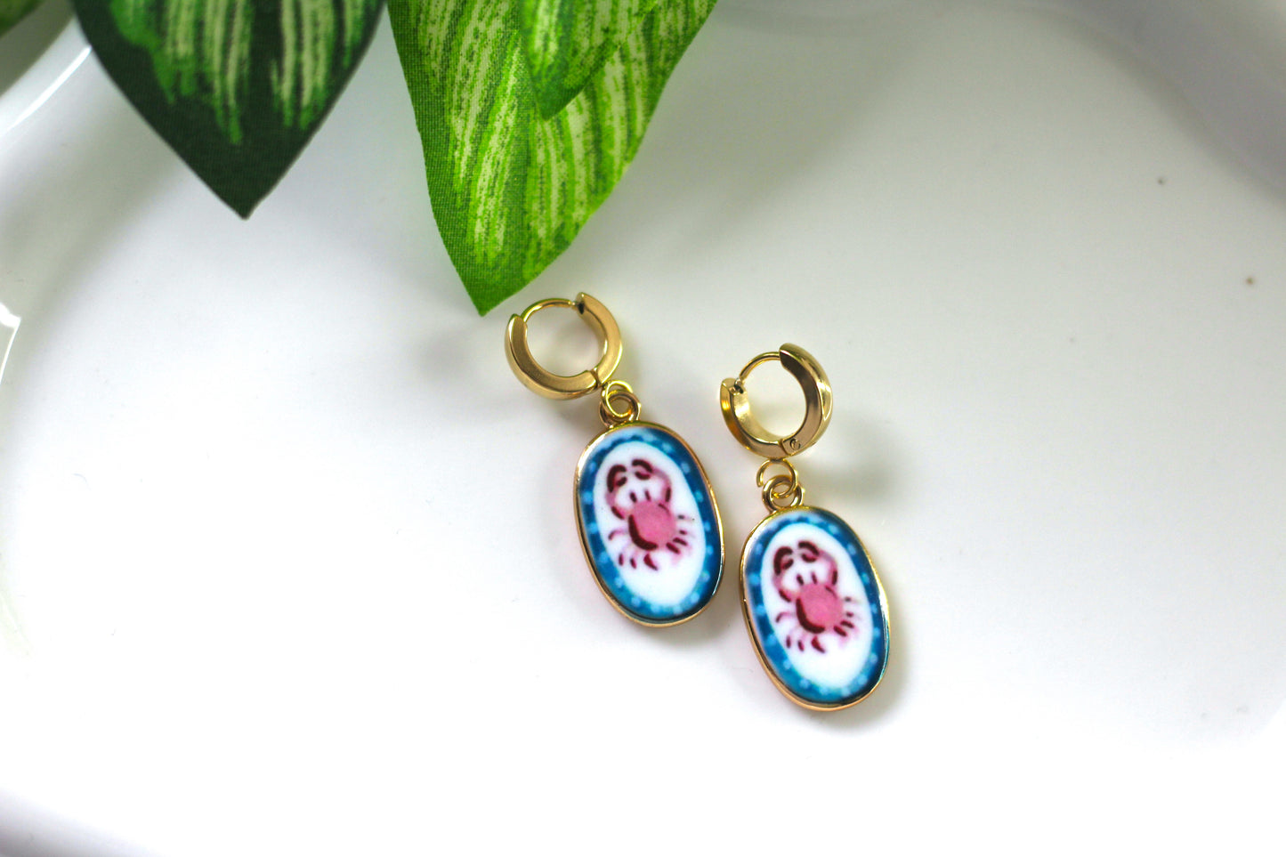 A pair of gold hoop earrings with oval enamel charms featuring a pink crab on a white background with blue floral borders, displayed on a white surface with a green leaf prop.