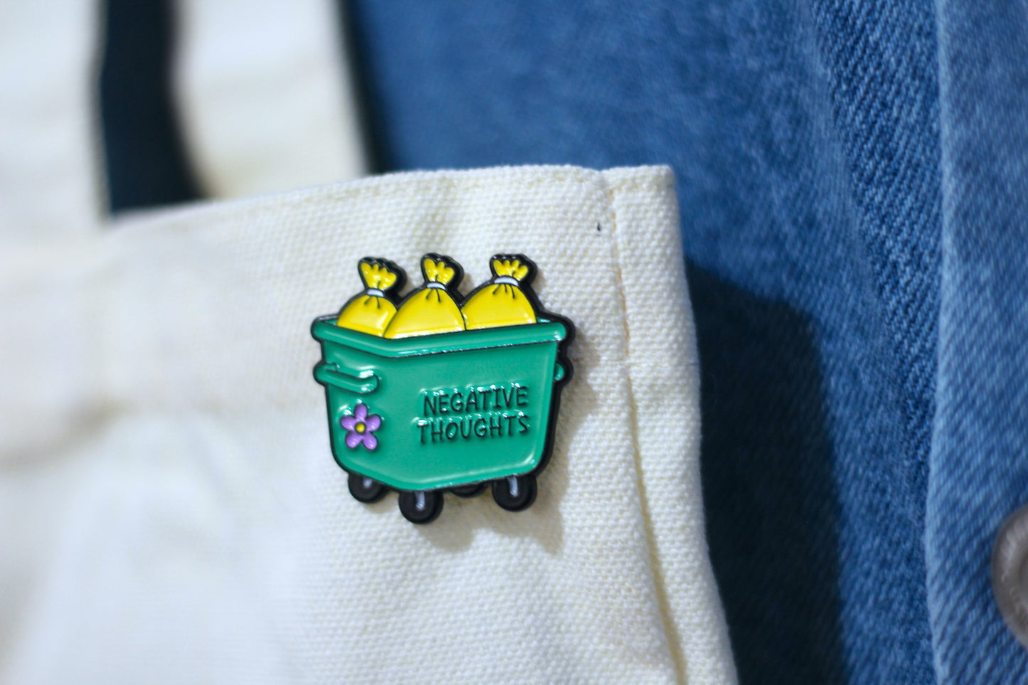 A close-up of a green enamel pin shaped like a trash can with three yellow bags of garbage and the words 'NEGATIVE THOUGHTS' on the front, pinned onto a white canvas tote next to a blue denim jacket.