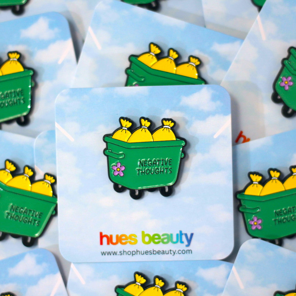 A green enamel pin shaped like a dumpster filled with yellow bags, with the words 'NEGATIVE THOUGHTS' and a small pink flower, displayed on a blue sky background. Multiple cards with the same pin are visible, and the brand name 'hues beauty' appears at the bottom.