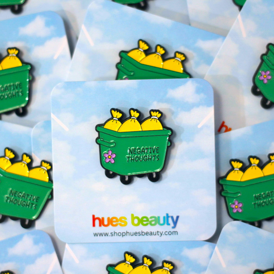 A green enamel pin shaped like a dumpster filled with yellow bags, with the words 'NEGATIVE THOUGHTS' and a small pink flower, displayed on a blue sky background. Multiple cards with the same pin are visible, and the brand name 'hues beauty' appears at the bottom.