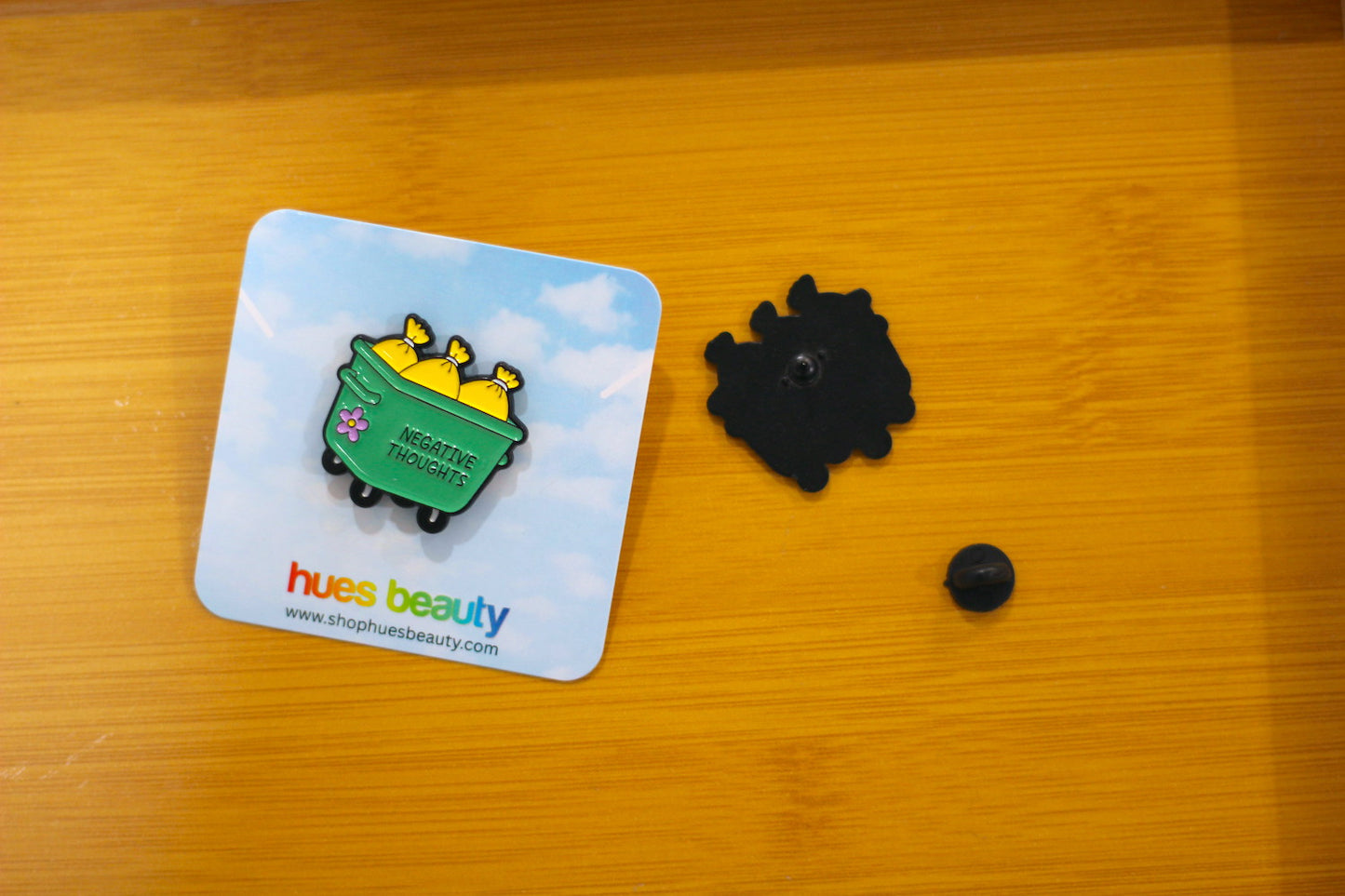 A small enamel pin shaped like a green dumpster filled with yellow trash cans, displayed on its branded backing card next to its black plastic clasp components on a wooden tabletop.