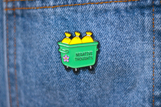 A green enamel pin shaped like a dumpster filled with yellow bags, with the words 'NEGATIVE THOUGHTS' and a small pink flower, shown pinned to blue denim.