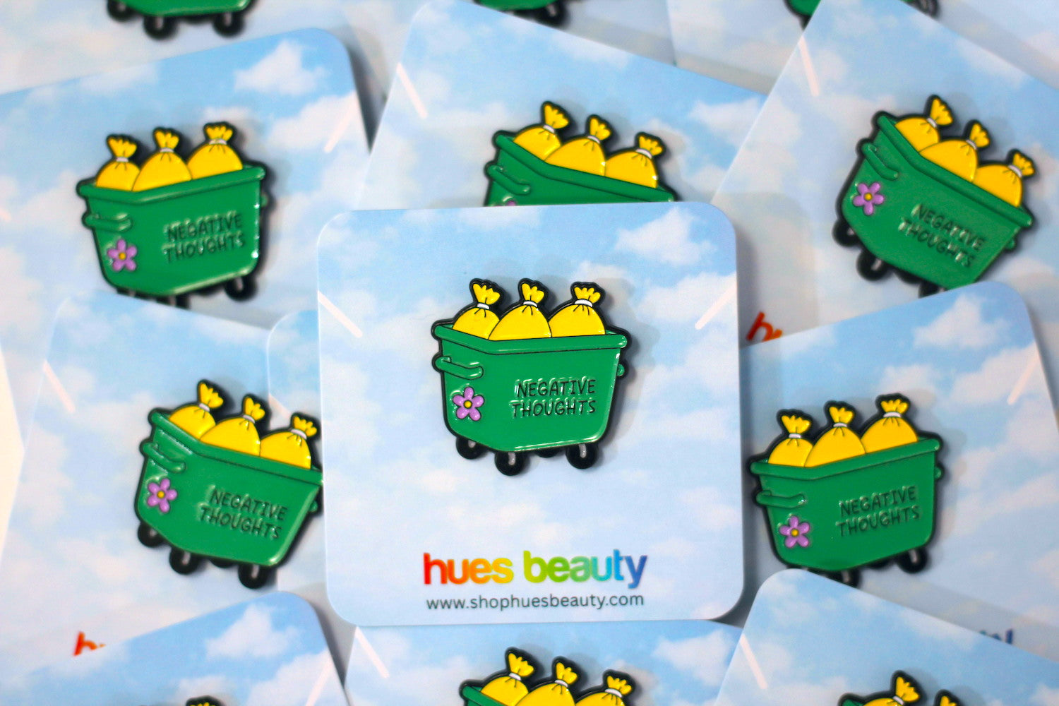 A close-up of several enamel pins displayed on matching backing cards. The pins feature a green dumpster filled with yellow bags, with the words 'NEGATIVE THOUGHTS' and small pink flower details.
