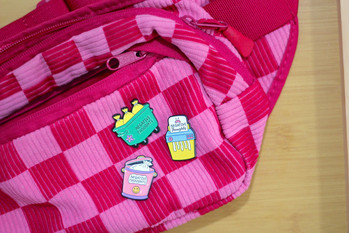 A close-up of a pink and magenta checkered corduroy bag with three colorful enamel pins attached to the front, set against a light wood background.