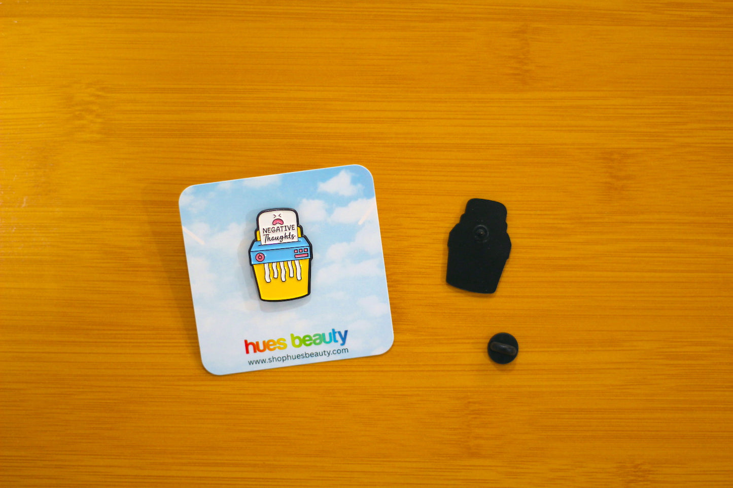 A small enamel pin shaped like a yellow shredder with a blue and white cloud background, shown next to its black plastic backing and a small black round piece on a warm wood surface. The card reads 'hues beauty'.