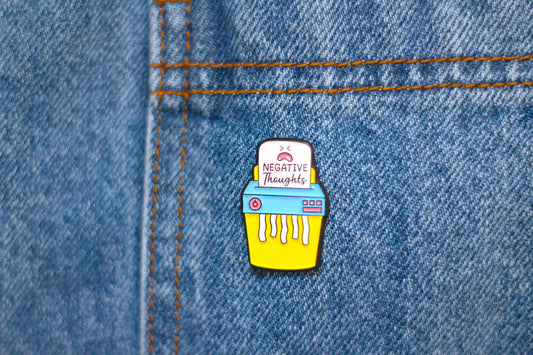 A small enamel pin shaped like a yellow shredder and a white sign reading 'NEGATIVE THOUGHTS' is pinned onto blue denim fabric.