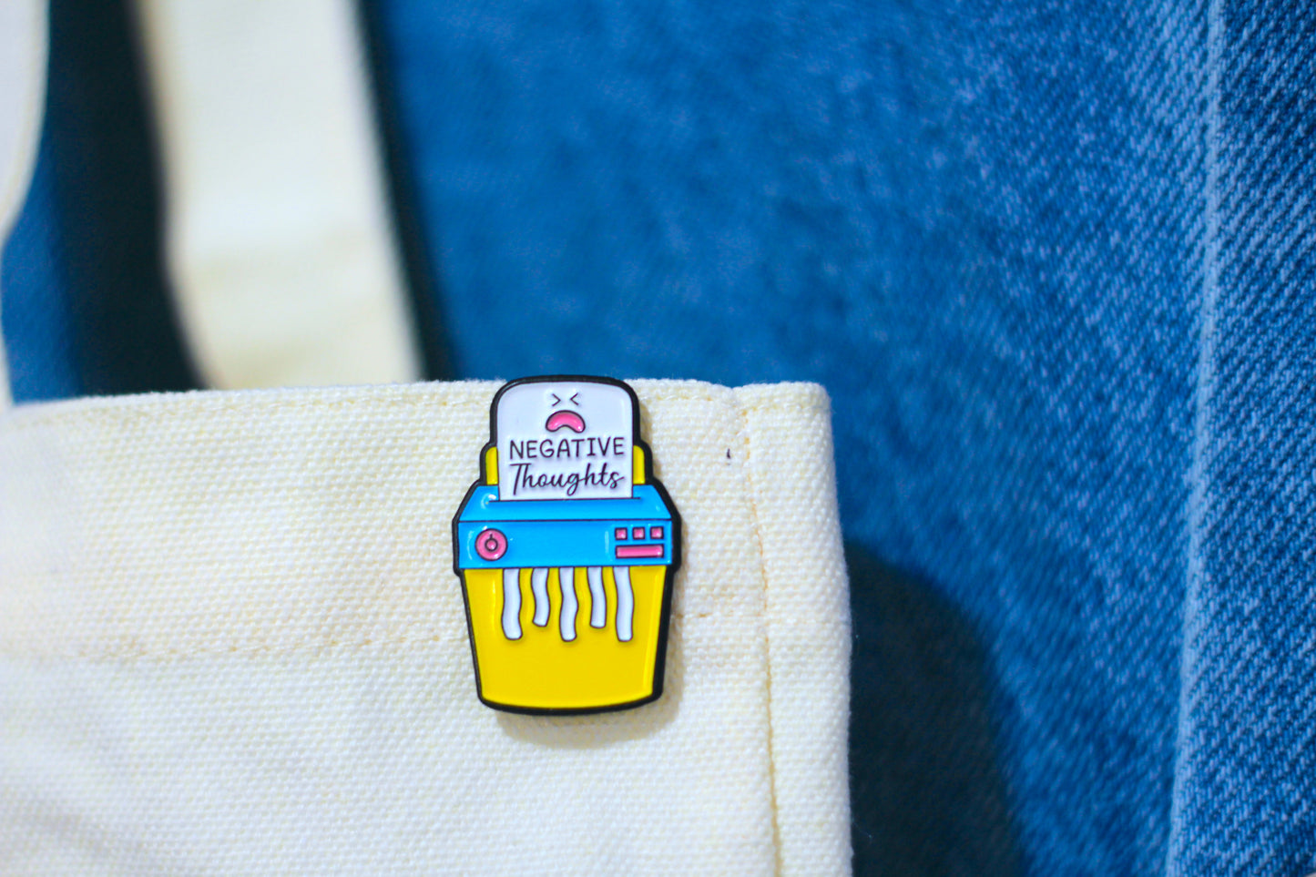 A close-up of a colorful enamel pin shaped like a shredder with a white sign reading 'NEGATIVE THOUGHTS', pinned to a cream canvas tote bag against a blue denim background.