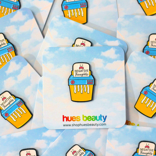 A set of enamel pin cards, featuring a light blue sky and white cloud pattern, each printed with a colorful shredder design and the brand name 'hues beauty'.