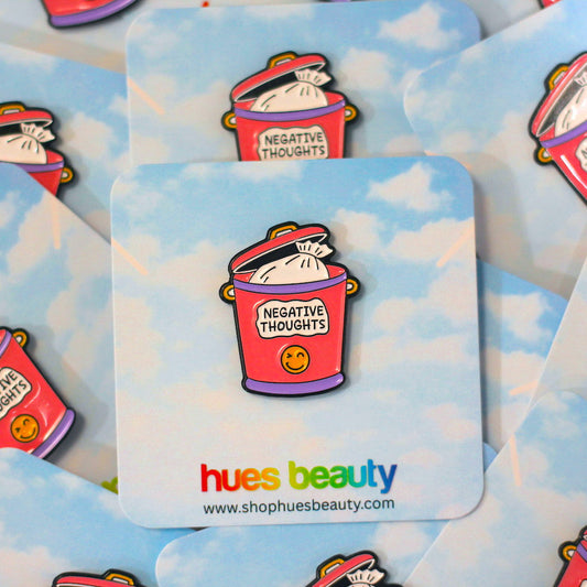 A close-up of several enamel pins arranged on a light blue card with a cloudy print. The pins show a pink trash can labeled 'NEGATIVE THOUGHTS' with a small yellow smiley face.