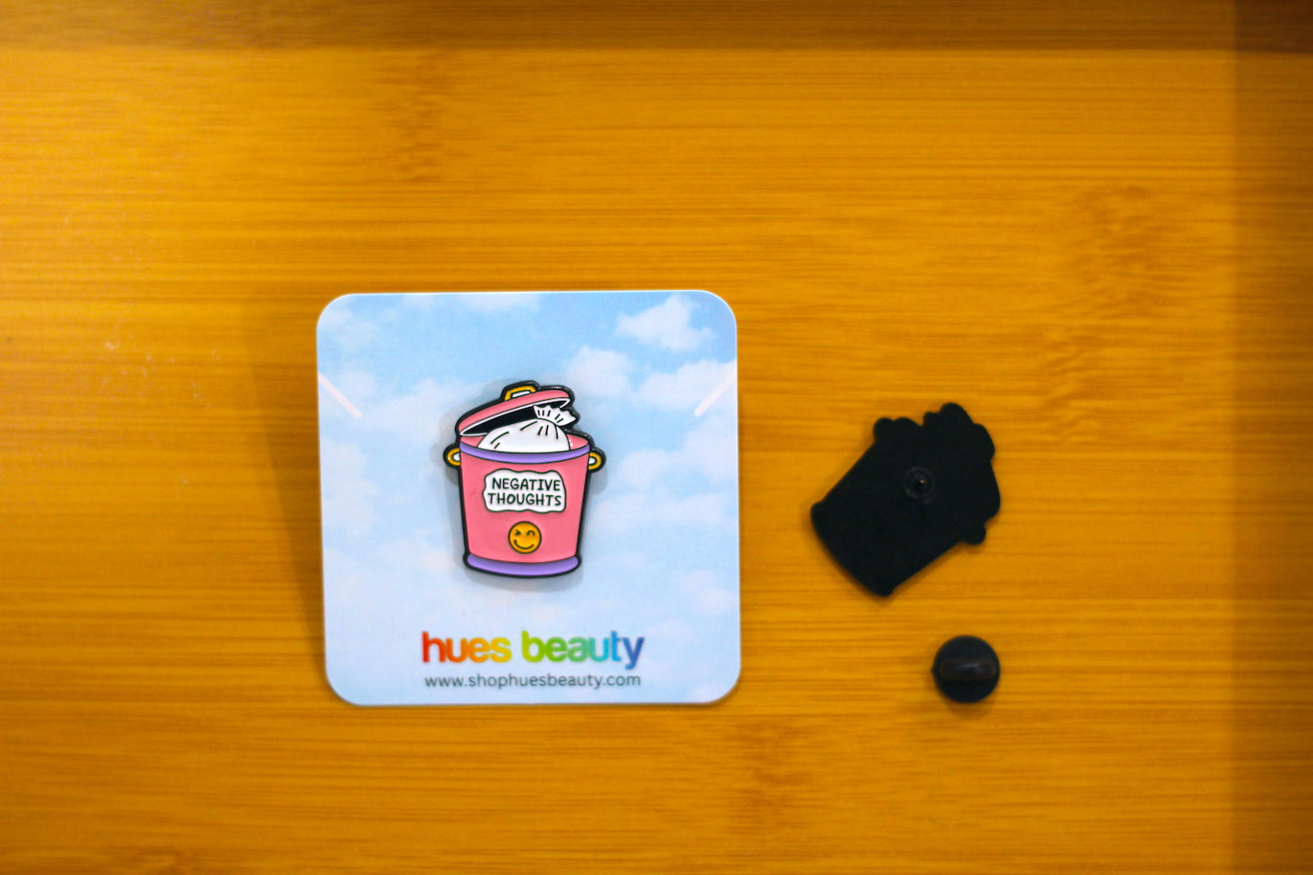 A square enamel pin displayed on a light blue backing card, placed on a wooden surface next to its black metal backing and small black fastener.