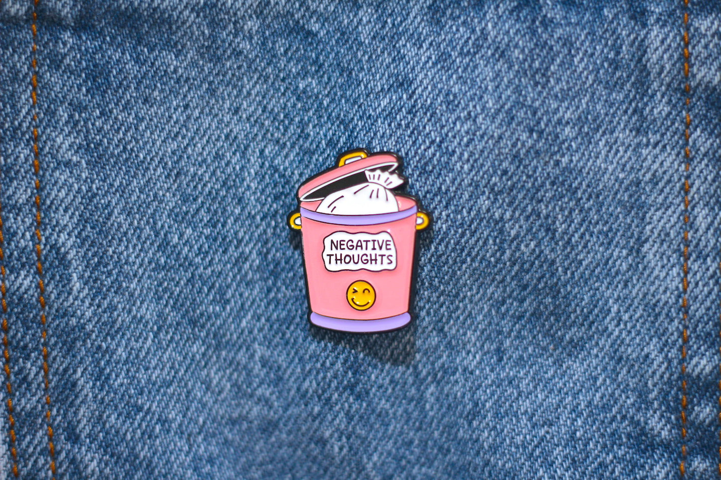 A small enamel pin shaped like a pink trash can with the words 'NEGATIVE THOUGHTS' and a yellow smiley face, placed on blue denim fabric.
