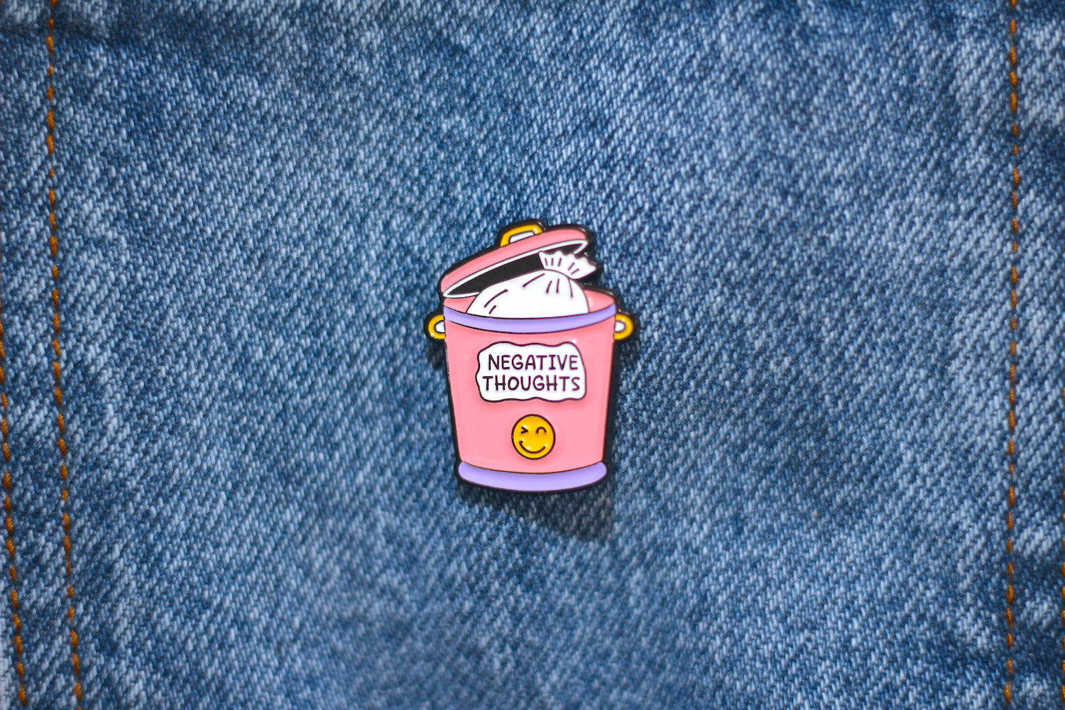 A small enamel pin shaped like a pink trash can with the words 'NEGATIVE THOUGHTS' and a yellow smiley face, placed on blue denim fabric.