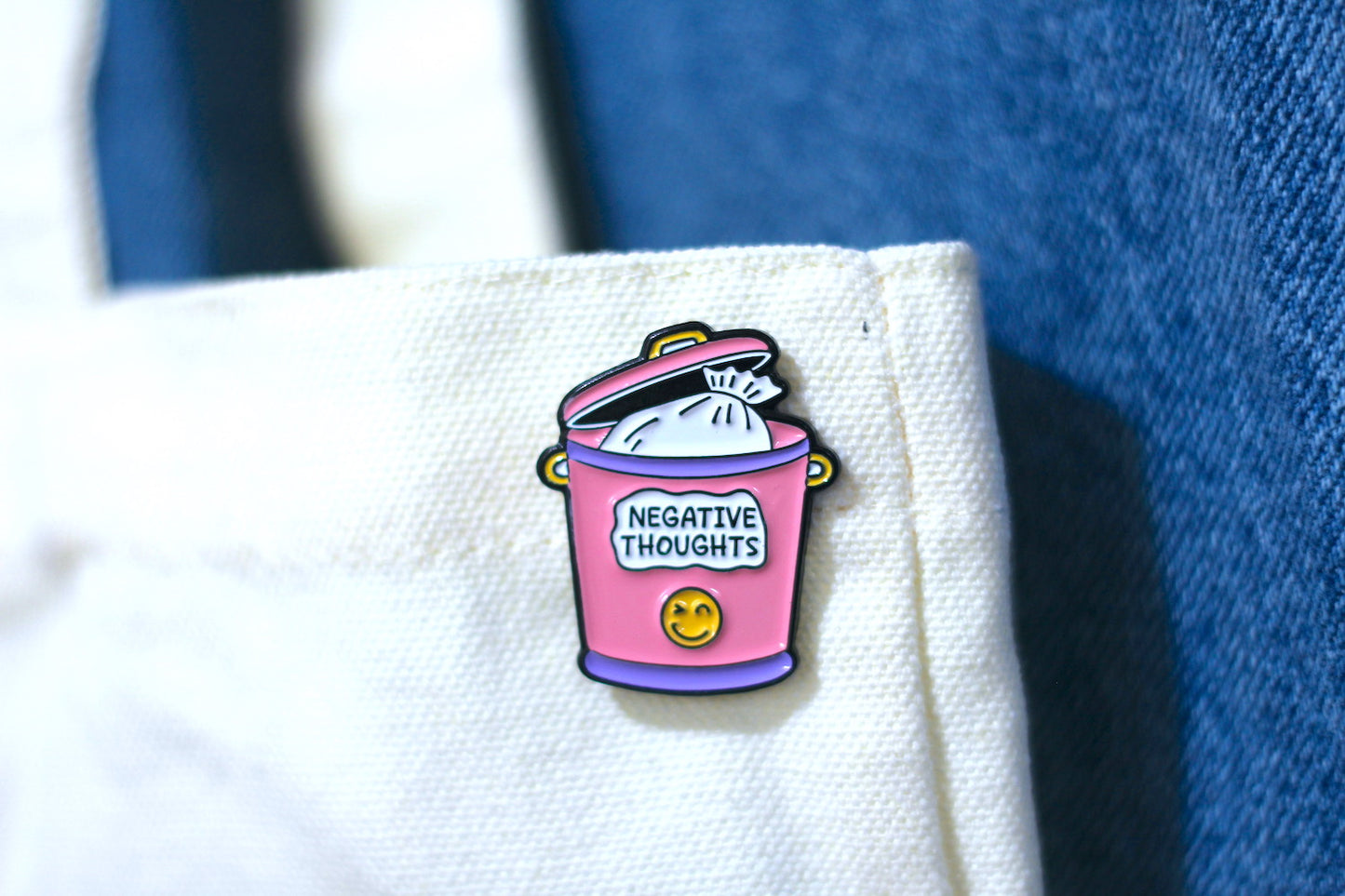 A small enamel pin shaped like a pink trash can with the words "NEGATIVE THOUGHTS" and a yellow smiley face, attached to a cream tote, with blue denim in the background.