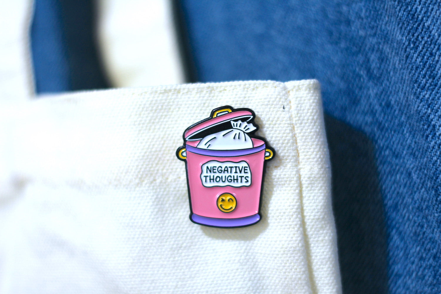 A small enamel pin shaped like a pink trash can with the words "NEGATIVE THOUGHTS" and a yellow smiley face, attached to a cream tote, with blue denim in the background.
