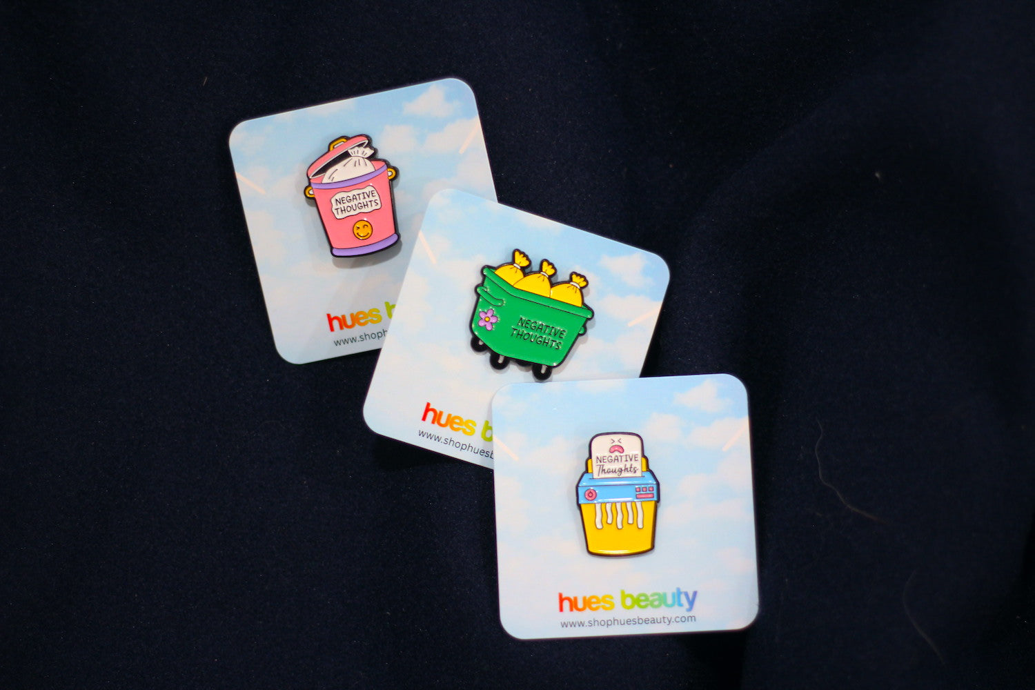 Three enamel pin badges with cloud backing cards, displayed on a dark fabric background. The badges feature colorful cartoon designs: a pink trash can, a green dumpster, and a yellow shredder, each with the brand name 'hues beauty' printed on the card.