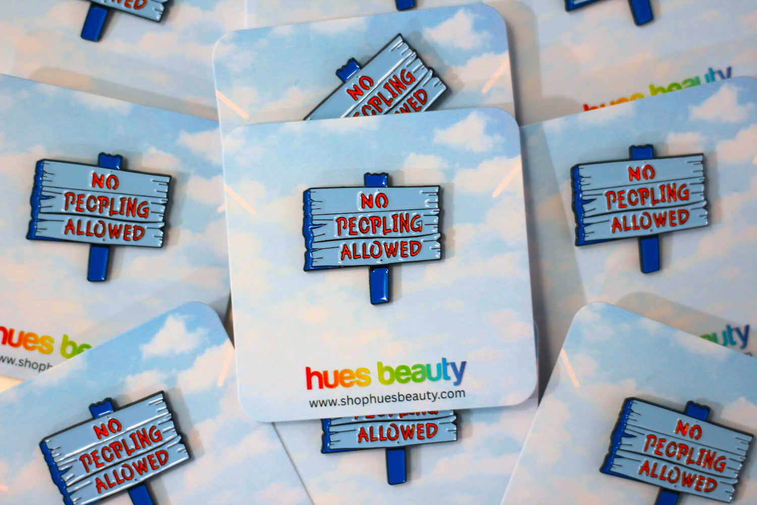 Several small enamel pins on backing cards, each featuring a wooden sign that reads “NO PEOPLING ALLOWED” in red lettering on a light blue-gray background, with blue sign posts. The cards are arranged on a matching sky-and-cloud print, and the center card shows the brand name “hues beauty” and website text.